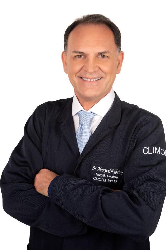 Dr. Maxwel Ribeiro sorrindo de braços cruzados, com o jaleco azul-marinho da Climor
